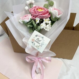 May include: A bouquet of crocheted flowers in pink and white, wrapped in white tissue paper and tied with a pink ribbon. The bouquet is in a brown cardboard box and has a card that says "Best wishes".