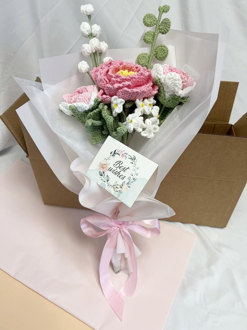 May include: A bouquet of crocheted flowers in pink and white, wrapped in white tissue paper and tied with a pink ribbon. The bouquet is in a brown cardboard box and has a card that says "Best wishes".