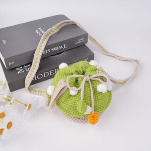 May include: A green crocheted drawstring bag with white pom-poms and a beige strap. The bag has a small orange pom-pom on the bottom.