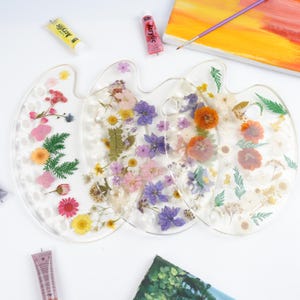 May include: Three clear resin paint palettes, each embedded with colorful dried flowers. Yellow and pink acrylic paint tubes are visible, along with a canvas and paintbrush, suggesting an art theme. The palettes are arranged on a white surface.