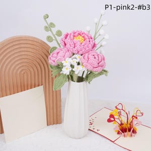 May include: A bouquet of pink and white crocheted flowers in a white vase. The flowers are arranged in a vase with a textured surface. The vase is sitting on a wooden surface. The flowers are made of yarn and are very realistic.