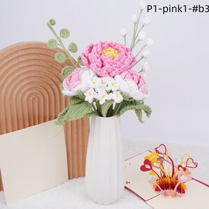 May include: A bouquet of crocheted flowers in a white vase. The flowers are pink, white, and green. The bouquet is sitting on a wooden surface. The text "P1-pink1-#b3" is visible in the upper right corner of the image.