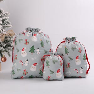 May include: Three gray drawstring gift bags with a festive Christmas pattern. The pattern includes Santa faces, Christmas trees, snowmen, and candy canes. The bags have red drawstrings and are in varying sizes.