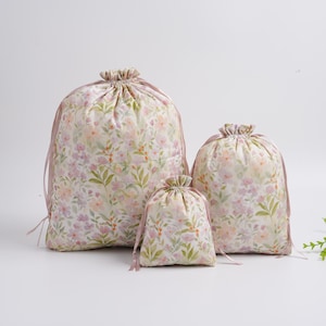 May include: Three drawstring bags of varying sizes, featuring a floral pattern in soft pastel colors. The bags have a cream background with pink, purple, and green floral accents. Each bag has a gathered top and matching pink drawstring closure.