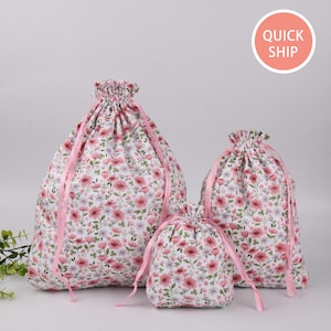May include: Three pink floral drawstring bags of different sizes. The bags are made of cotton fabric with a pink floral pattern. The bags have pink drawstrings.