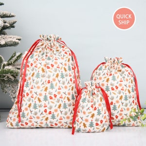 May include: Three white fabric gift bags with red ribbon ties. The bags are decorated with a Christmas pattern of gingerbread men, Christmas trees, and holly.