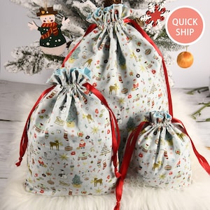 May include: Set of three Christmas gift bags with a festive print of Christmas trees, reindeer, snowmen, and other holiday icons. The bags are made of a light blue fabric with red ribbon ties.