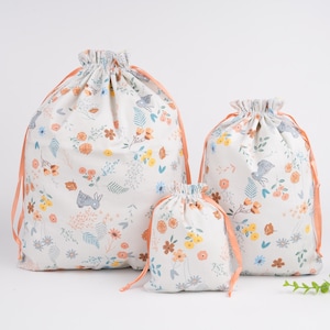 May include: Three drawstring bags in varying sizes, featuring a white background with a floral and bunny print in shades of orange, yellow, blue, and green. The bags have orange drawstrings.