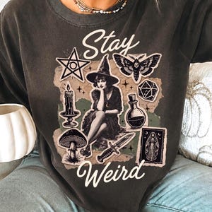 May include: Dark gray sweatshirt with the words "Stay Weird" and various witchy graphics, including a pentagram, witch, moth, mushroom, and dagger. The design is printed on a comfortable, casual top.