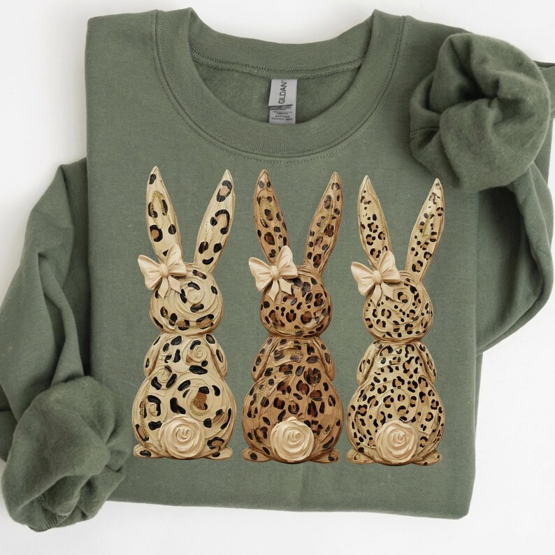 May include: An olive green sweatshirt featuring three decorative bunny figures. The bunnies have a leopard print design with cream-colored bows and a rose detail on the back. The sweatshirt is made of a soft material.