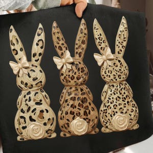 Może przedstawiać: Trzy dekoracyjne figurki królików z wzorem w cętki. Każdy królik ma kremową kokardę i ogon w kształcie róży. Króliki są w kolorze beżowym, brązowym i kremowym, na czarnym tle.