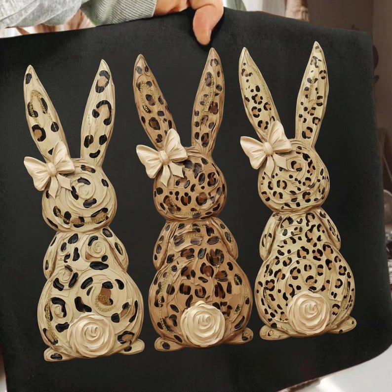 May include: Three decorative bunny figurines with a leopard print design. Each bunny has a cream-colored bow and a rose-shaped tail. The bunnies are tan, brown, and cream-colored, set against a black background.