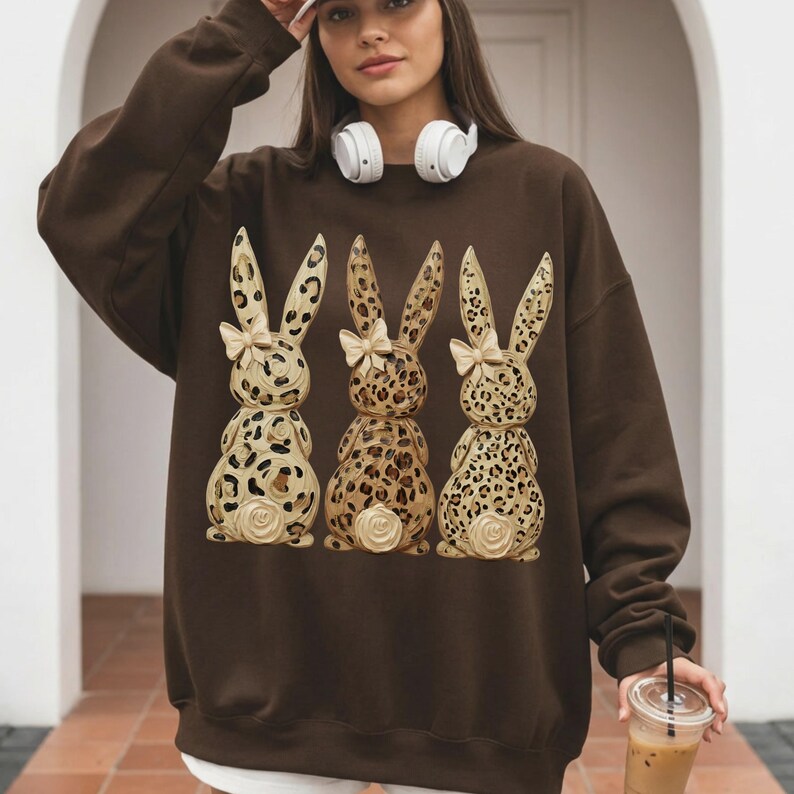 May include: A brown sweatshirt featuring three decorative Easter bunny figures. The bunnies have a leopard print design with cream-coloured bows and floral accents. The sweatshirt is a solid brown colour.