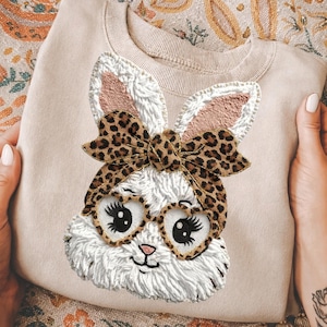 May include: A beige sweatshirt featuring an embroidered bunny face with a leopard print bow and heart-shaped glasses. The bunny has white fur, pink inner ears, and a small black nose and whiskers. The sweatshirt is held by hands.