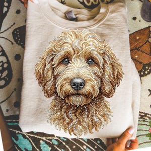 May include: Beige sweatshirt featuring a detailed, jeweled portrait of a dog's face. The dog's fur is rendered in shades of brown and gold, with sparkling accents. The dog has a black nose and dark eyes. The sweatshirt is folded.
