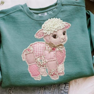 May include: Teal-colored sweatshirt featuring a patchwork lamb design. The lamb is primarily pink, with various patterned patches including floral, polka dots, and gingham. The lamb has a fluffy white wool head and a small bow. The sweatshirt has a crew neck.