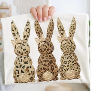 May include: A white fabric featuring three decorative bunny figures. Each bunny has a leopard print pattern in shades of brown and tan, with a cream-colored bow and a rose detail at the base. The bunnies are arranged in a row.