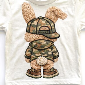 Pu&ograve; includere: T-shirt bianco con un design ricamato di un coniglietto con cappellino da baseball, giacca e scarpe mimetica. Il coniglietto ha una soffice pelliccia marrone e una piccola coda bianca. Il design &egrave; dettagliato e strutturato.