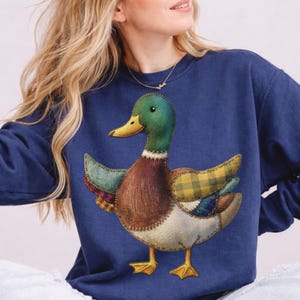 May include: Navy blue sweatshirt with a patchwork duck design. The duck has a green head, brown body, and wings with plaid and patterned fabric. The duck's beak is yellow, and the feet are yellow. The sweatshirt is a solid navy blue colour.