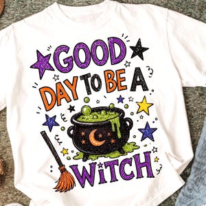 Op de afbeelding: Wit T-shirt met een kleurrijke Halloween-afbeelding. Het ontwerp bevat de tekst "GOOD DAY TO BE A WITCH" in paars, oranje en zwart, samen met een ketel, sterren en een bezem.