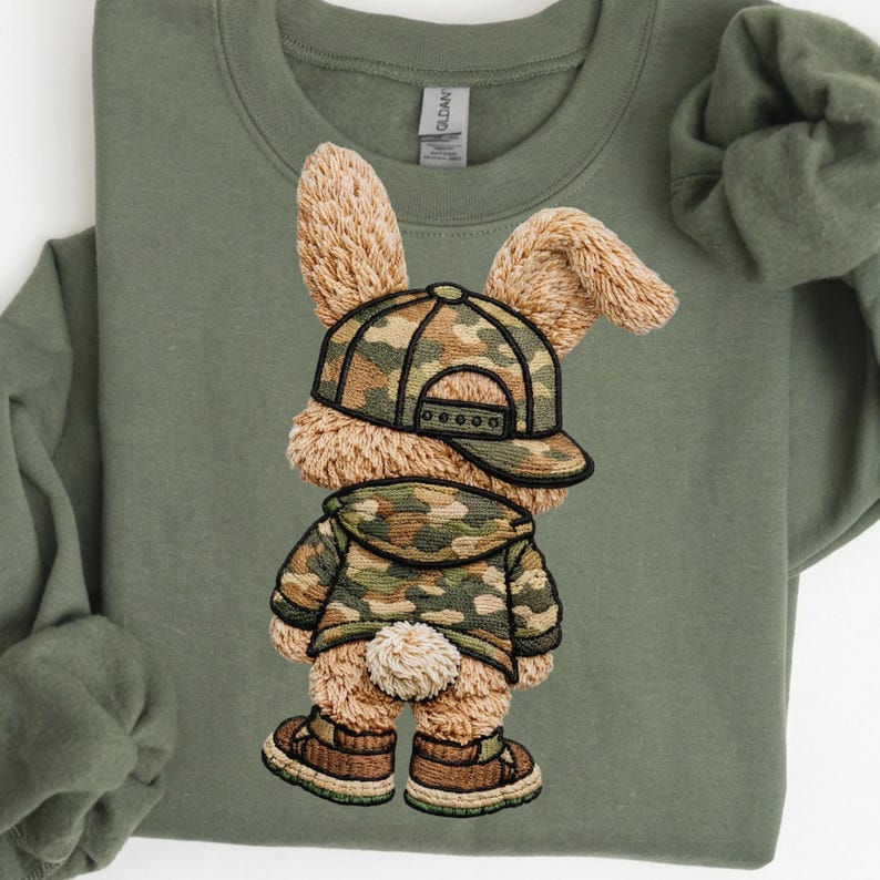 Pu&ograve; includere: Felpa verde oliva con un coniglio ricamato che indossa un berretto da baseball mimetico, una giacca e scarpe. Il coniglio ha una pelliccia soffice e strutturata e una piccola coda bianca. Il design &egrave; centrato sulla felpa.