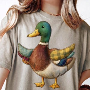 May include: A light gray t-shirt featuring a patchwork duck design. The duck has a green head, brown body, and wings with plaid and patterned fabric. The duck's beak and feet are yellow, and the design is stitched onto the shirt.