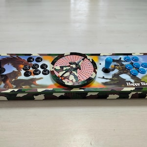 Op de afbeelding: Aangepaste arcade game controller met een camouflage ontwerp. Het heeft een zwarte joystick en knoppen aan de linkerkant, een centraal cirkelvormig ontwerp en een blauwe joystick en knoppen aan de rechterkant. De controller heeft een "Happy Birthday" bericht.