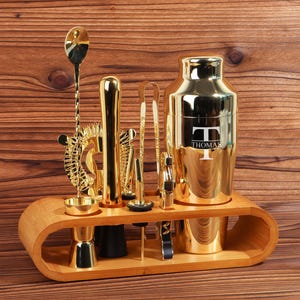 Pode incluir: Um conjunto de ferramentas de bar douradas com um shaker, um jigger, um coador, uma colher e outras ferramentas. As ferramentas são exibidas em um suporte de madeira. O shaker tem o nome "Thomas" gravado.