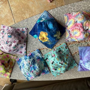May include: A collection of colorful fabric bowl cozies. Designs include Hello Kitty, Pikachu, Mickey Mouse, Snoopy, and tie-dye patterns. These square cozies are designed to hold bowls and protect hands from heat.