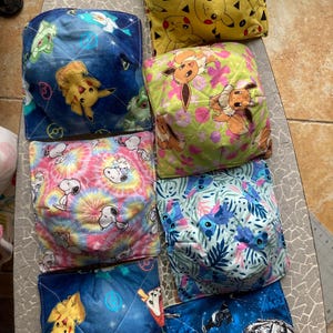 May include: Eight square-shaped fabric bean bags with different cartoon character designs. The bean bags are made of quilted fabric and have a variety of colors, including blue, green, yellow, pink, and white. The designs include Pokemon characters, Snoopy, and Stitch.