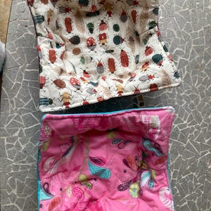 May include: Two quilted fabric bowl cozies. The top one is white with a pattern of red and green bugs. The bottom one is pink with a paisley pattern.