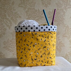 May include: A yellow fabric storage bin with a black and white polka dot trim. The bin is filled with yarn and knitting needles. The bin has a pattern of scissors, yarn balls, and other sewing-related items.