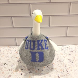 Peut inclure: Figurine d'oie blanche avec un bec jaune et un pull gris. Le pull porte le mot "DUKE" en lettres bleues. L'oie a des ailes blanches et se trouve sur une surface mouchetée.