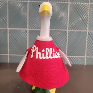 Op de afbeelding: Een keramieken ganzenbeeldje met een witte kop en nek, gele snavel en een rode vilten cape. Op de cape staat het woord "Phillies" in witte letters. De basis is groen en geel. Het beeldje is een sport-thema verzamelobject.