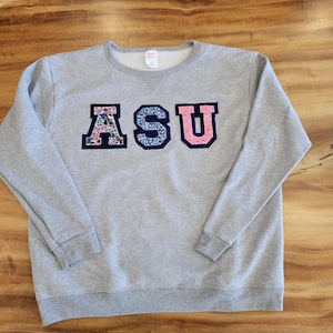Op de afbeelding: Een grijs sweatshirt met de letters "ASU" in grote, kleurrijke, gepatroneerde letters.