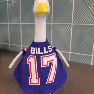 Op de afbeelding: Een wit ganzenbeeldje dat een blauw vilten shirt draagt met het woord BILLS en het nummer 17 in het wit met rode omlijning op de voorkant gedrukt. De gans heeft een gele snavel en zwarte ogen, en het shirt heeft kleine witte mouwtjes.