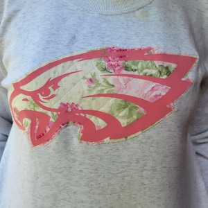 May include: A gray sweatshirt with a pink floral eagle design. The eagle is outlined in pink and the floral design is a patchwork of pink and green flowers on a cream background.