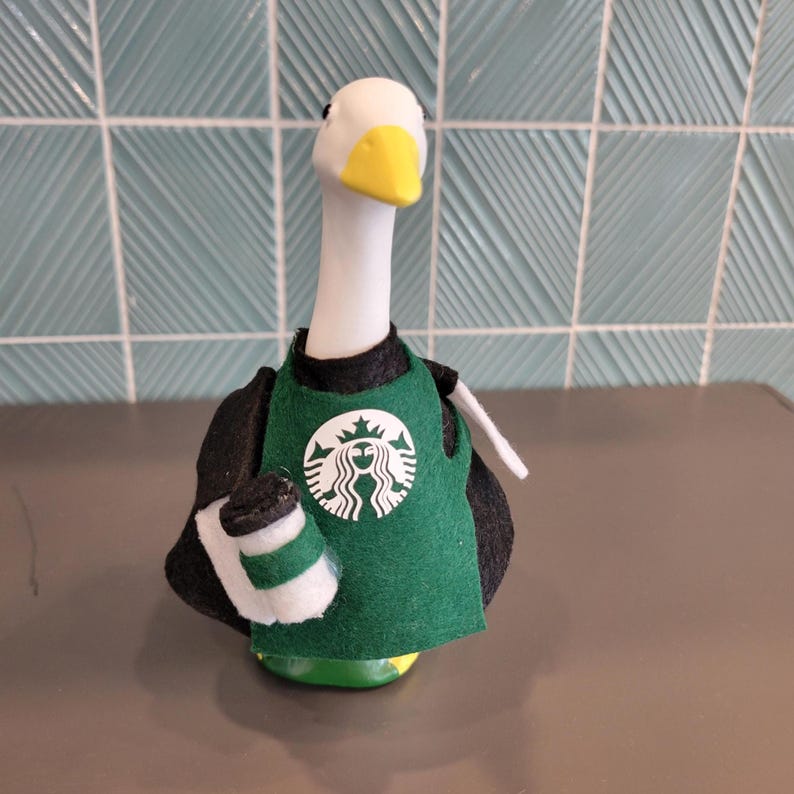 May include: A whimsical goose figurine wearing a green barista apron with the Starbucks logo and holding a coffee cup. The goose has a white head, yellow beak, and black and white body. The figurine is on a gray surface with a blue tile background.