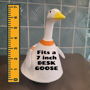 May include: A white desk goose figurine with a yellow beak and orange accents. The text "Fits a 7 inch DESK GOOSE" is printed on the front. A yellow ruler is placed next to the goose.