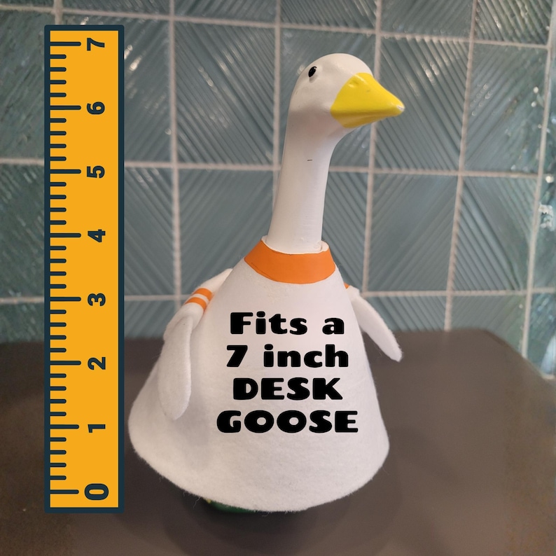 May include: A white desk goose figurine with a yellow beak and orange accents. The text "Fits a 7 inch DESK GOOSE" is printed on the front. A yellow ruler is placed next to the goose.