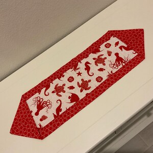 Könnte beinhalten: Ein roter und weißer Tischläufer mit einem Meereslebewesen-Design. Der Läufer zeigt rote Seepferdchen, Seesterne, Muscheln und Kraken auf weißem Hintergrund. Der Läufer hat einen roten Rand mit weißen Punkten.