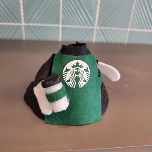 May include: A felt doll wearing a green apron with the Starbucks logo and holding a small coffee cup. The doll is black and white, with a white wing. The background is a light teal tile.