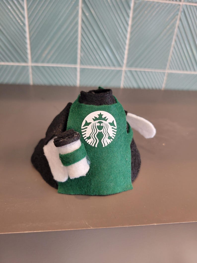 May include: A felt doll wearing a green apron with the Starbucks logo and holding a small coffee cup. The doll is black and white, with a white wing. The background is a light teal tile.