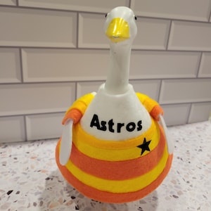 May include: A white goose figurine wearing a felt outfit with orange, yellow, and white stripes. The outfit has the word "Astros" in black and a black star. The goose has a yellow beak.