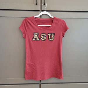 May include: A maroon t-shirt with the letters "ASU" across the chest. The letters are decorated with a floral pattern. The shirt is hanging on a white hanger.
