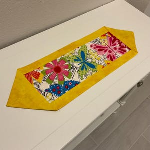 May include: A yellow table runner with a white background and a colorful floral and butterfly design. The runner has a yellow border.