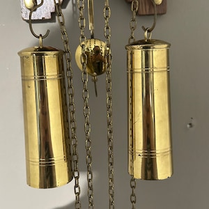 May include: Two gold-toned weights hang from chains attached to a wooden clock face. The weights are cylindrical and have a smooth, polished finish.