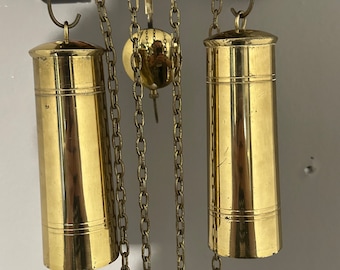 May include: Two gold-toned weights hang from chains attached to a wooden clock face. The weights are cylindrical and have a smooth, polished finish.