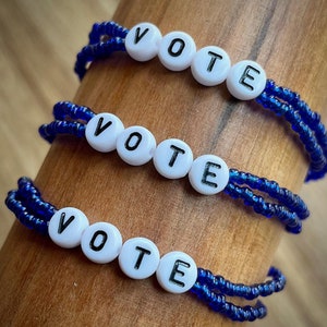 May include: Three blue beaded bracelets with white letter beads spelling "VOTE". The bracelets are displayed on a wooden surface. The beads are a deep blue color, and the letters are black.