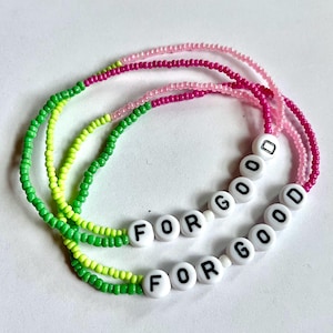 Könnte beinhalten: Drei übereinanderliegende Perlenarmbänder mit grünen, gelben und rosa Perlen. Die Armbänder buchstabieren "For Good" in weißen Buchstaben.